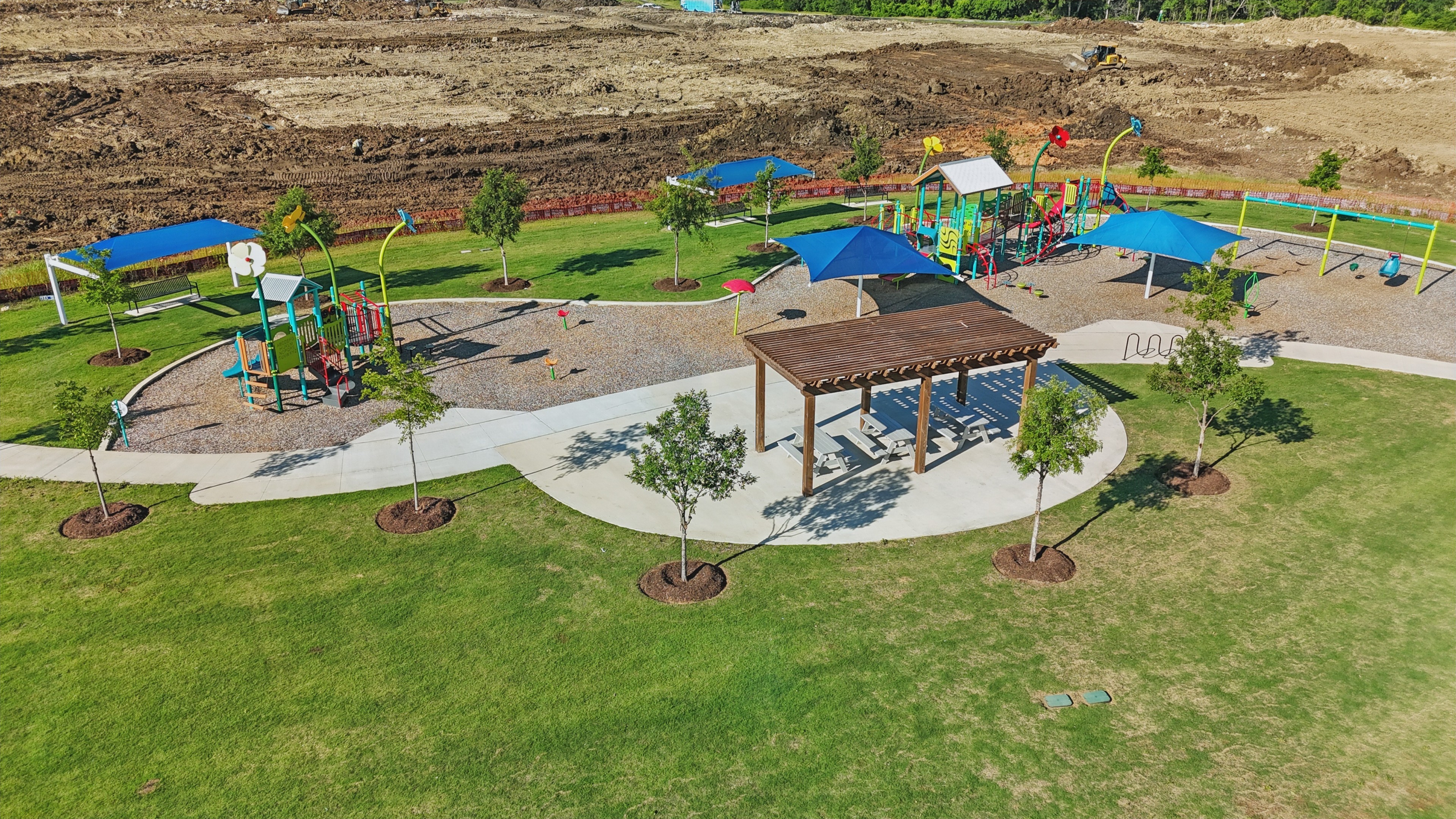 Playground in Wildflower Ranch with colorful structures, shaded tables, green lawn, and construction in the background.