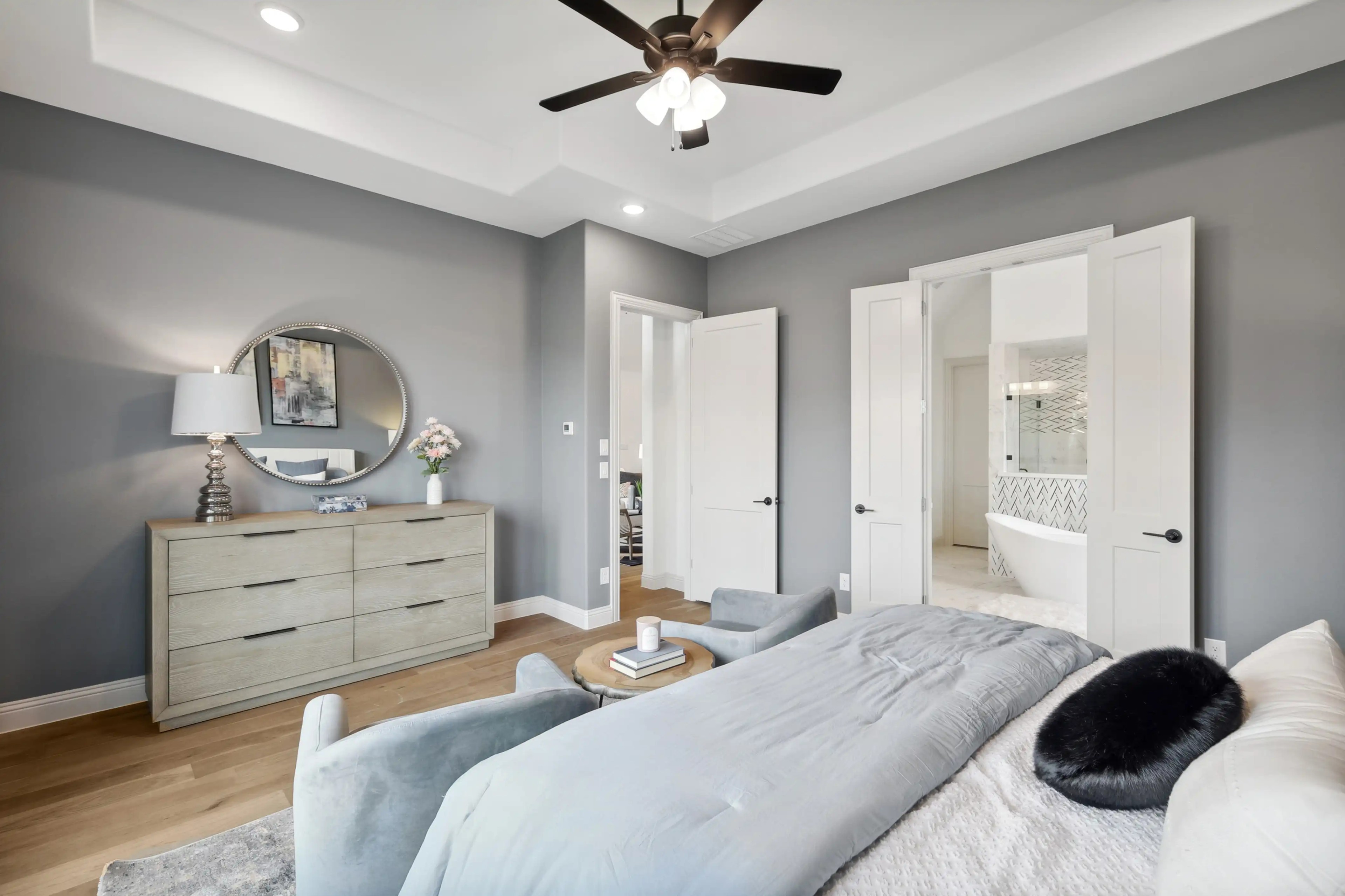 Elegant bedroom with modern decor, featuring a round mirror, light wood dresser, and en-suite bath in Westside Preserve.