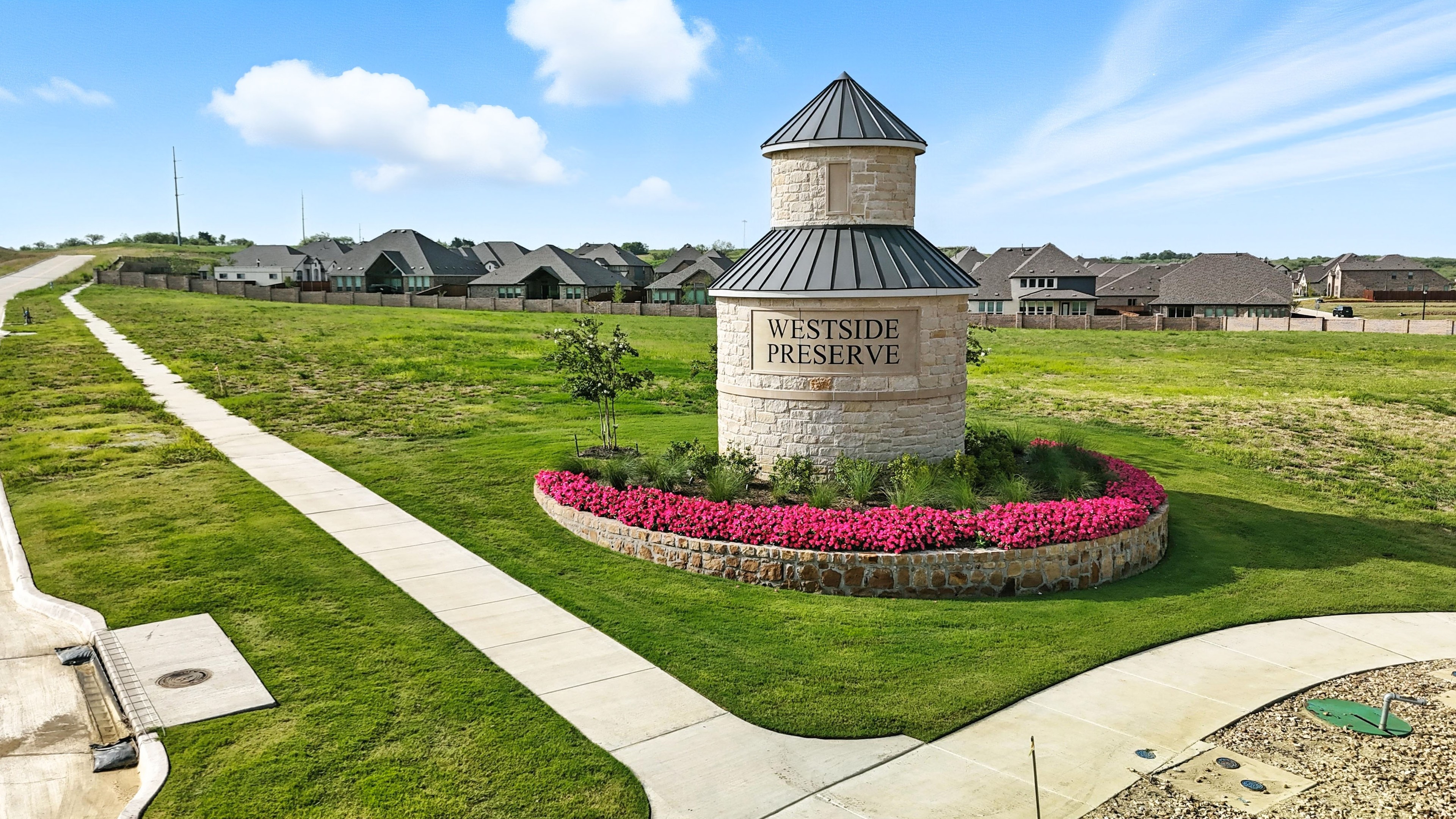 Stone tower with "Westside Preserve" sign, surrounded by vibrant flowers, near modern homes in green landscape.