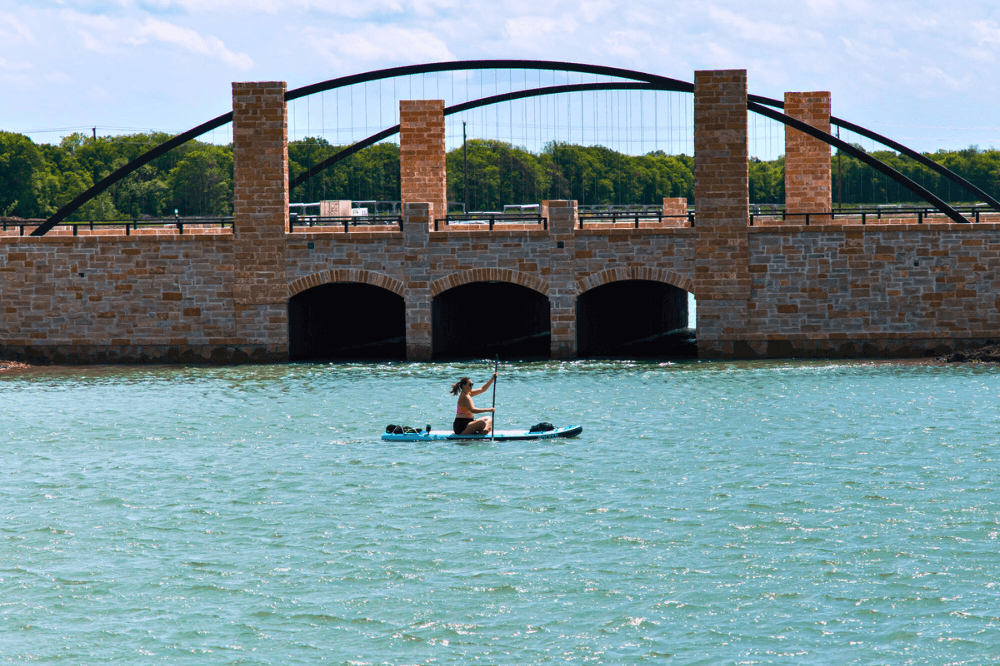 Stone bridge with arched supports over lake, paddleboarder in foreground, lush greenery, Solterra Texas community.