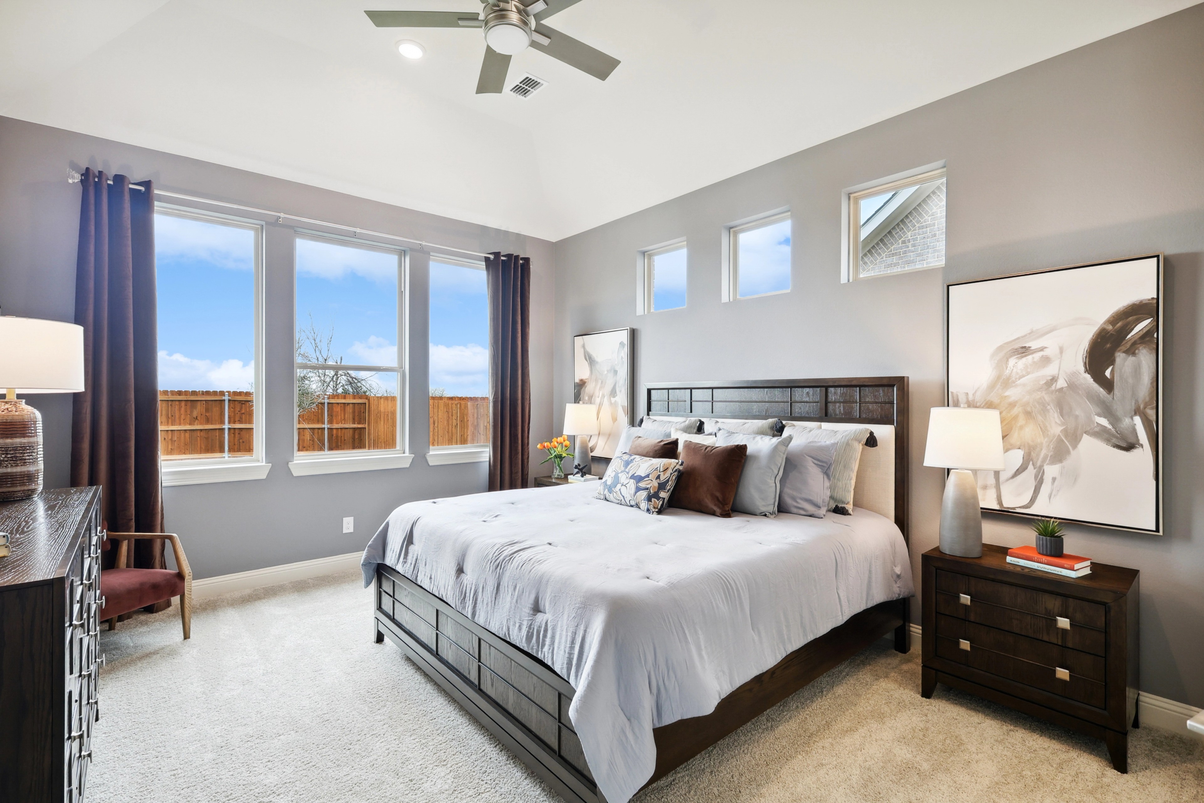 Luxurious bedroom in The Oaks with plush carpeting, elegant decor, and large windows. Modern ceiling fan enhances comfort.