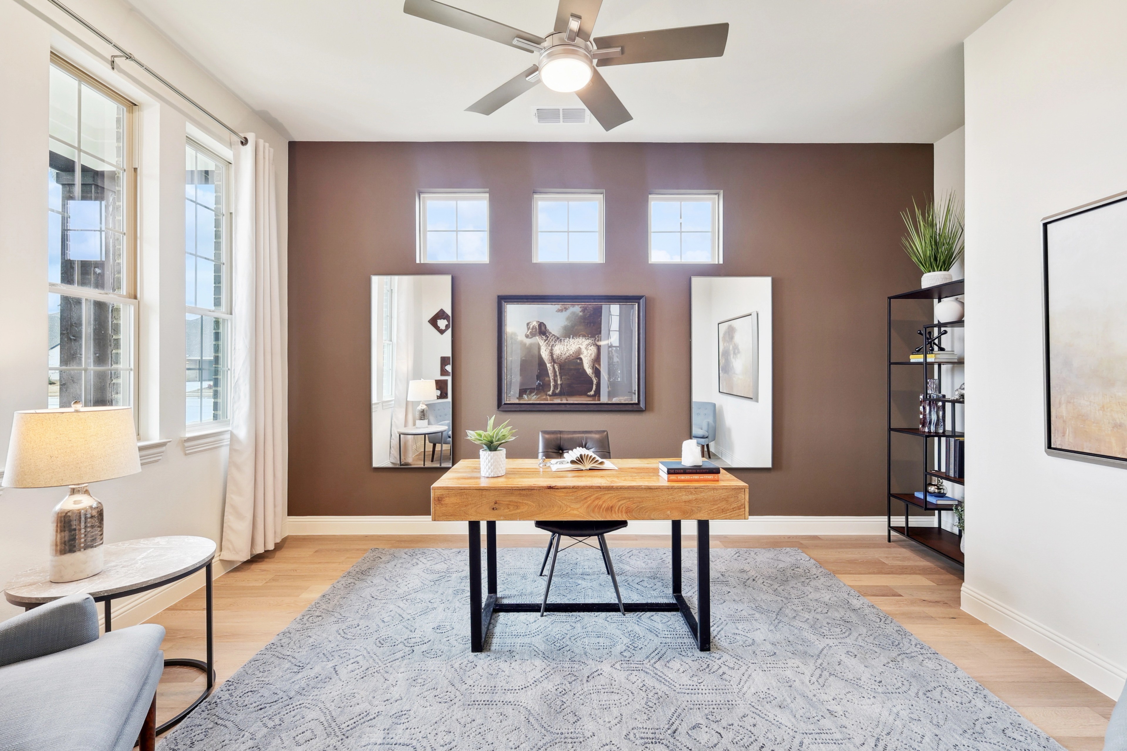 Modern home office with wood desk, abstract art, large windows, ceiling fan in The Oaks community.