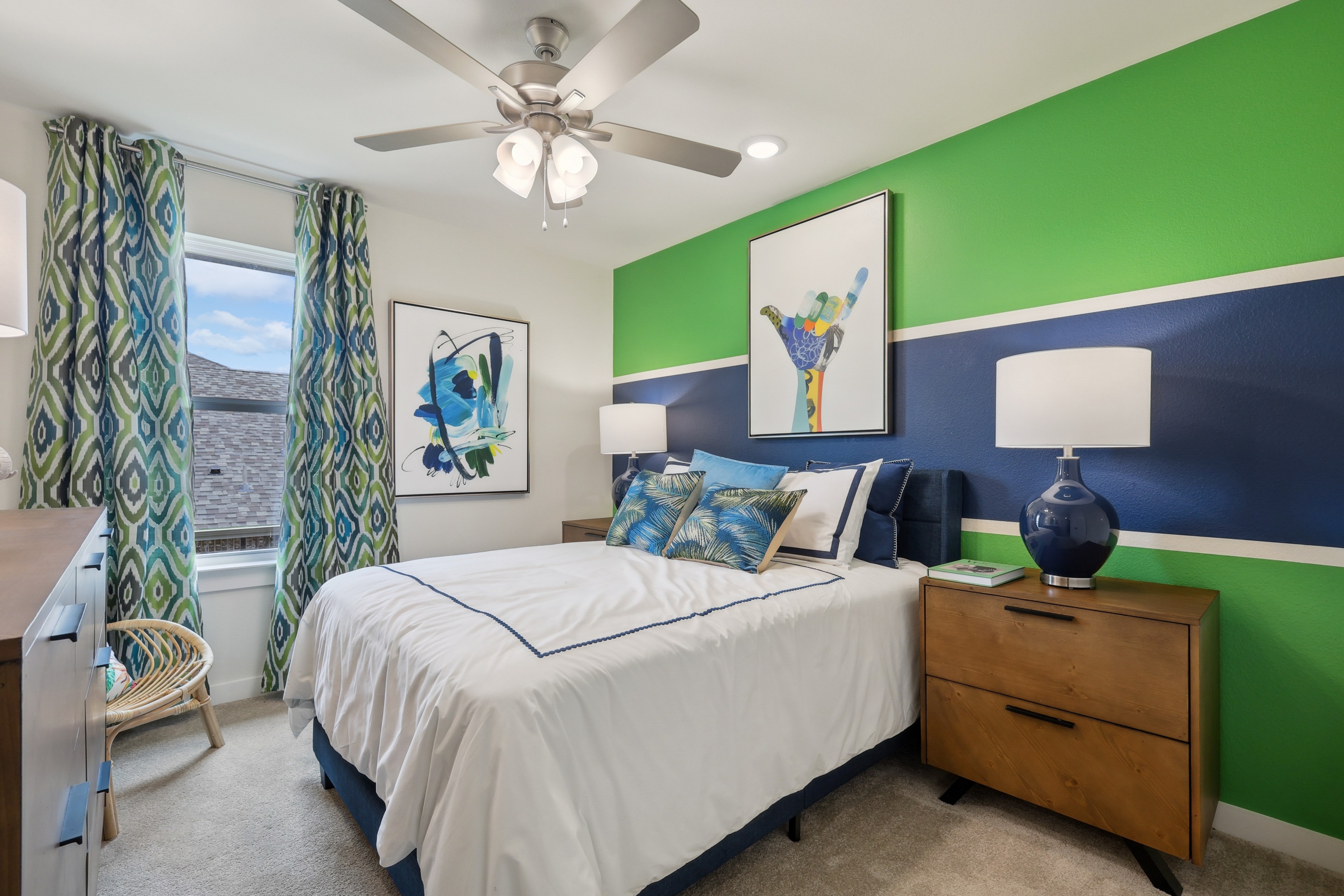 Modern bedroom with vibrant green and blue accent wall, abstract art, ceiling fan, and patterned curtains.