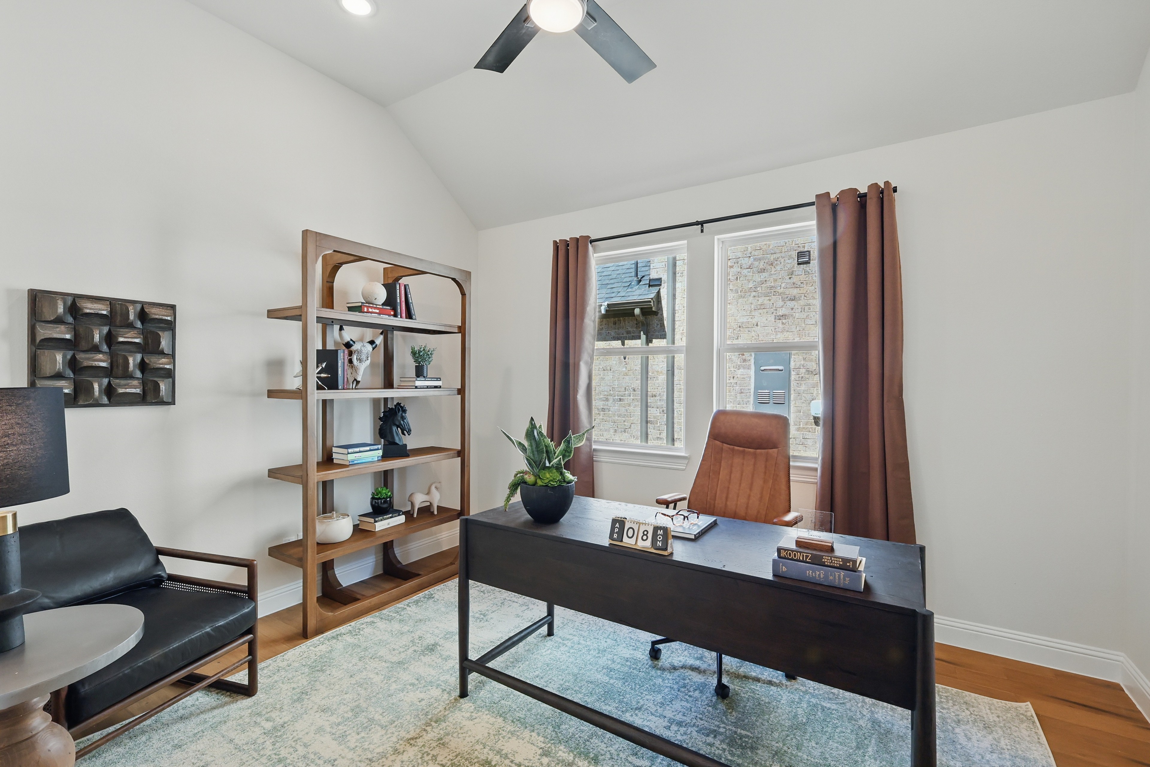 Modern home office with wood desk, leather chair, and shelf. Large window, vaulted ceiling. Green Oaks Preserve.