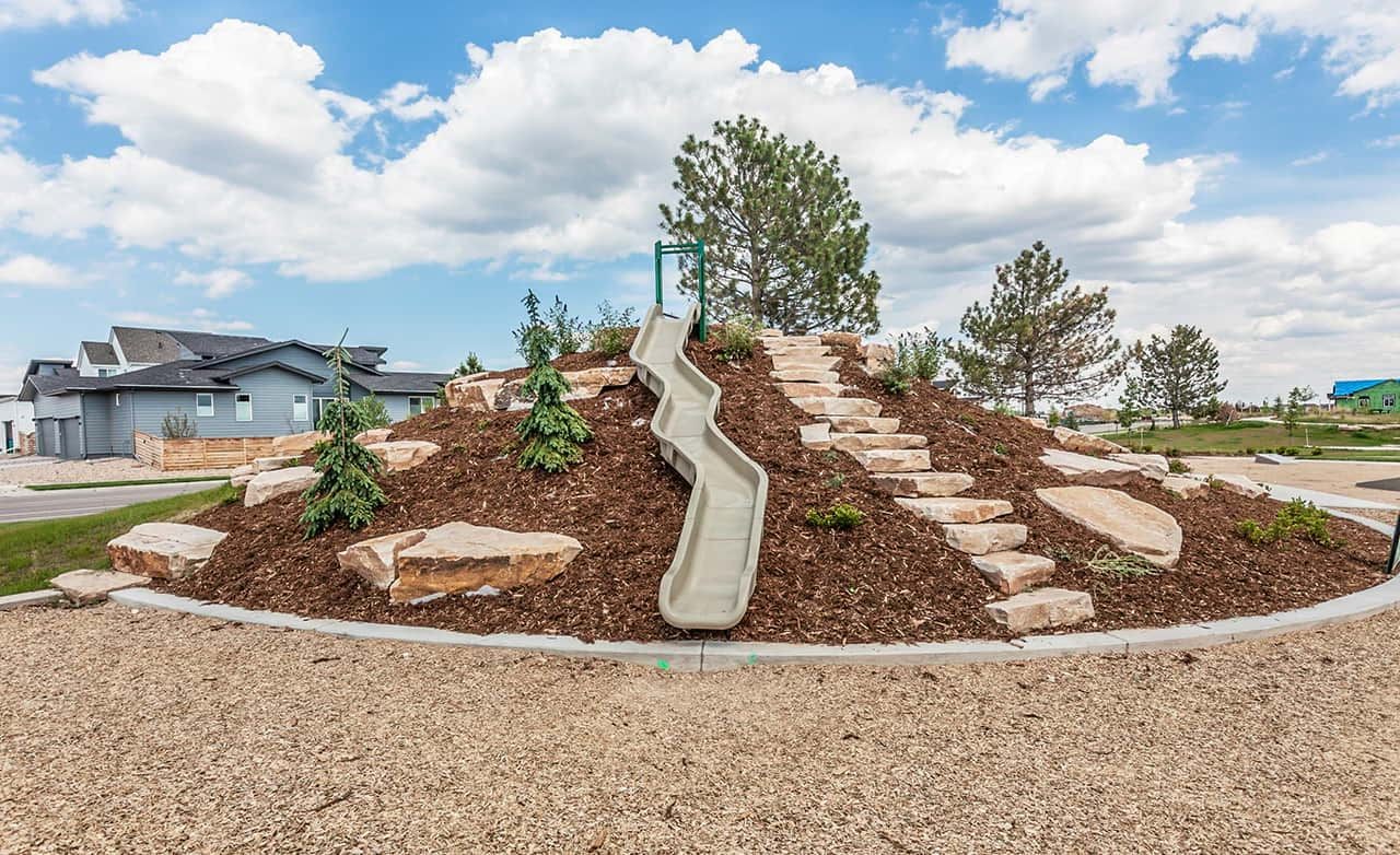 Modern playground with slide on landscaped hill, Trailside on Harmony, featuring boulders and lush greenery under blue skies.
